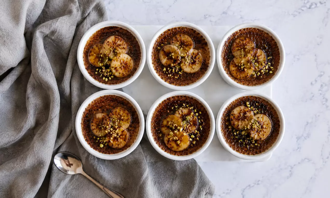 Schoko-Crème Brûlée mit dunkler, karamellisierter Kruste, belegt mit goldbraunen Honig-Bananenscheiben und gehackten Pistazien in weißen Förmchen