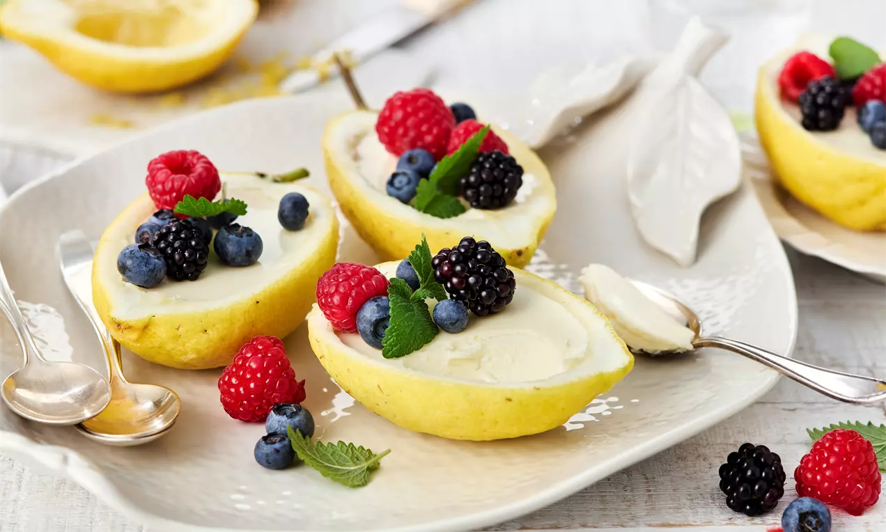 Helle Zitronenhälften gefüllt mit cremigem Dessert, garniert mit frischen roten und blauen Beeren sowie Minzblättern auf einem weißen Teller
