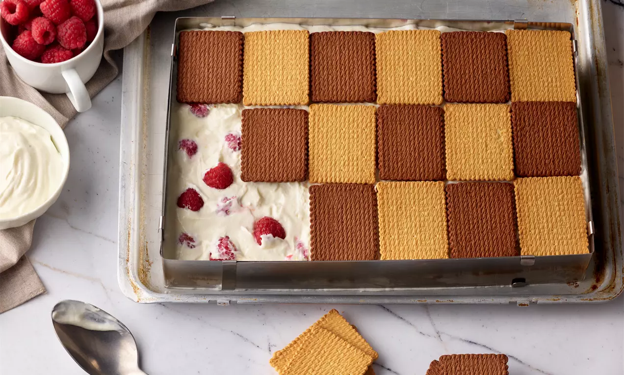 Keksschnitten mit hellbraunen und schokoladenbraunen Keksen schachbrettartig angeordnet auf Sahnecreme mit Himbeeren in einer eckigen Form