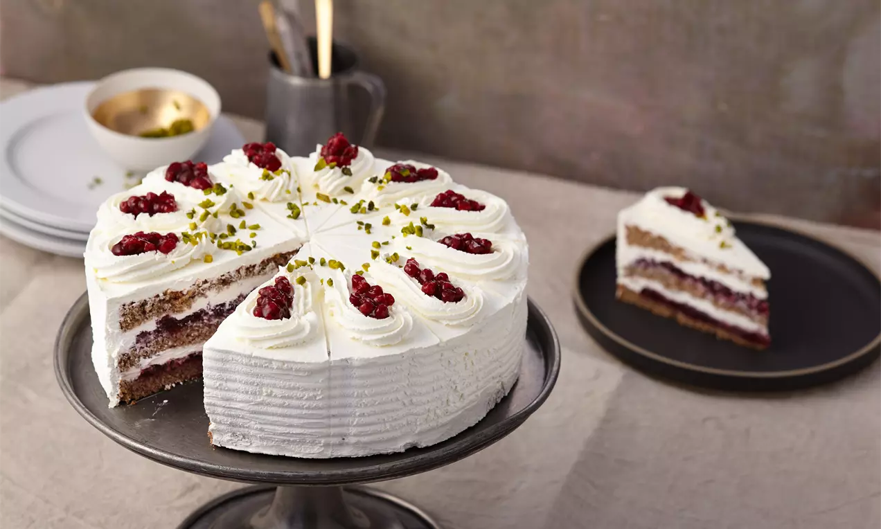 Lüneburger Buchweizentorte mit weißer Sahneverzierung, roten Preiselbeeren und grünen Pistazien auf dunkler Kuchenplatte angerichtet
