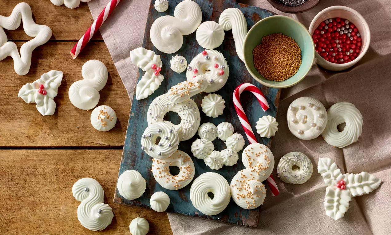 Schneeweiße Windbäckerei in spiralförmigen und sternförmigen Mustern auf rustikalem Brett mit bunten Streuseln und Zuckerstangen arrangiert