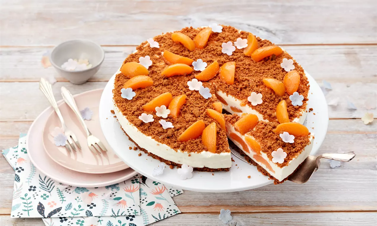 Käsekuchen ohne Backen mit weißer Cremefüllung, knusprigem Keksboden, Aprikosenstücken und Zuckerdekoren auf weißer Tortenplatte