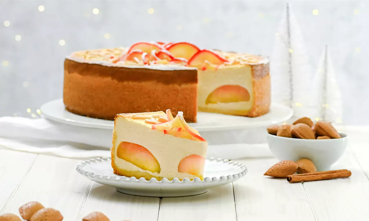 Bratapfel-Käsekuchen-Stück mit goldbraunem Boden und Apfelspalten, garniert mit Mandelstiften vor dem ganzen Kuchen auf weißem Teller