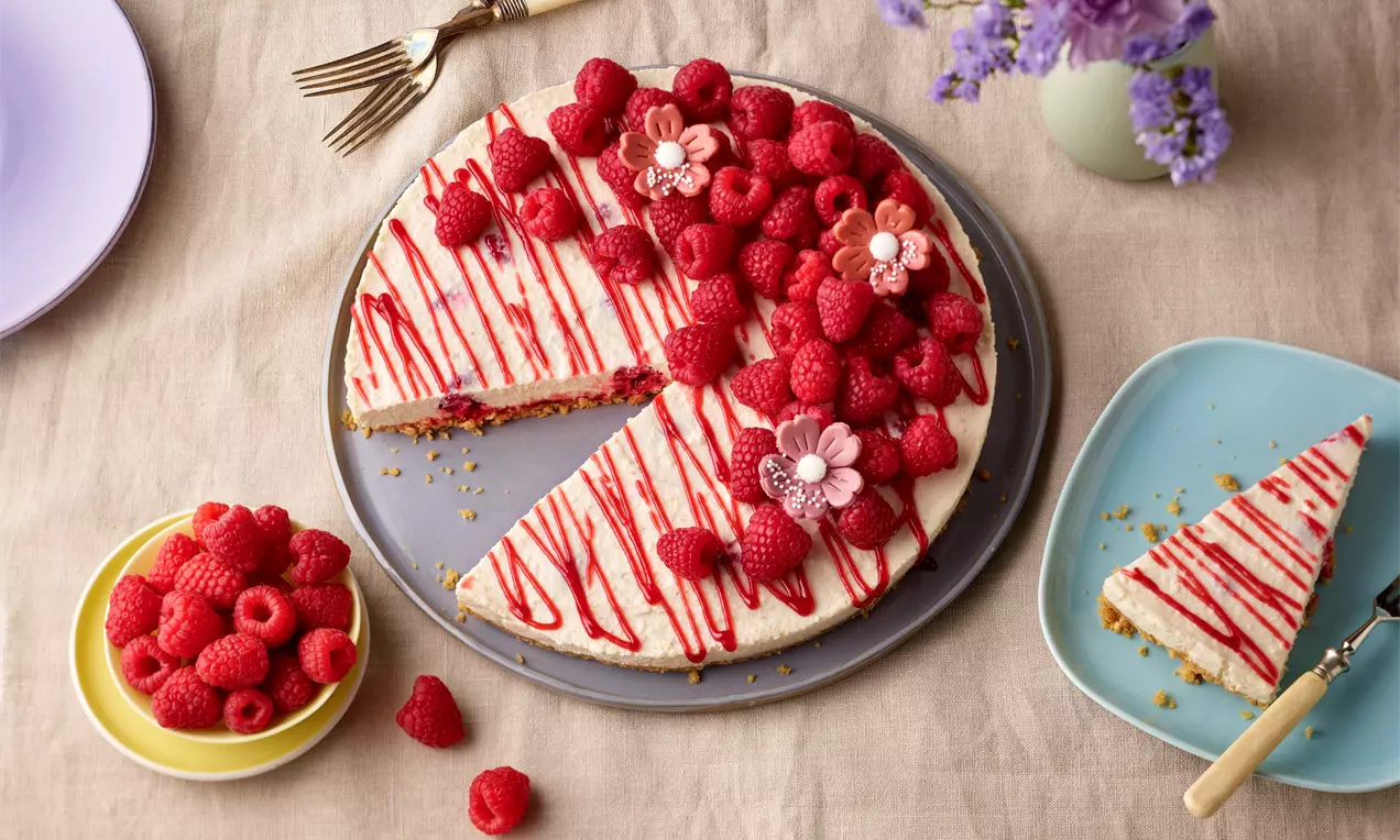 Veganer Cheesecake mit heller Füllung, knusprigem Boden, roten Himbeeren und Zuckerguss auf einem Teller, daneben Blumen und Geschirr