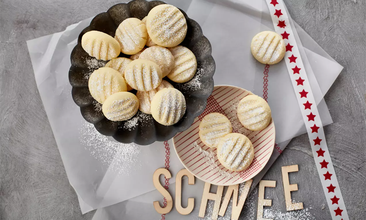 Zartweiße, runde Schneeflöckchen mit feinem Puderzucker auf dunklem Teller und hellem Streifen-Teller, dekoriert mit rotem Band und Buchstaben