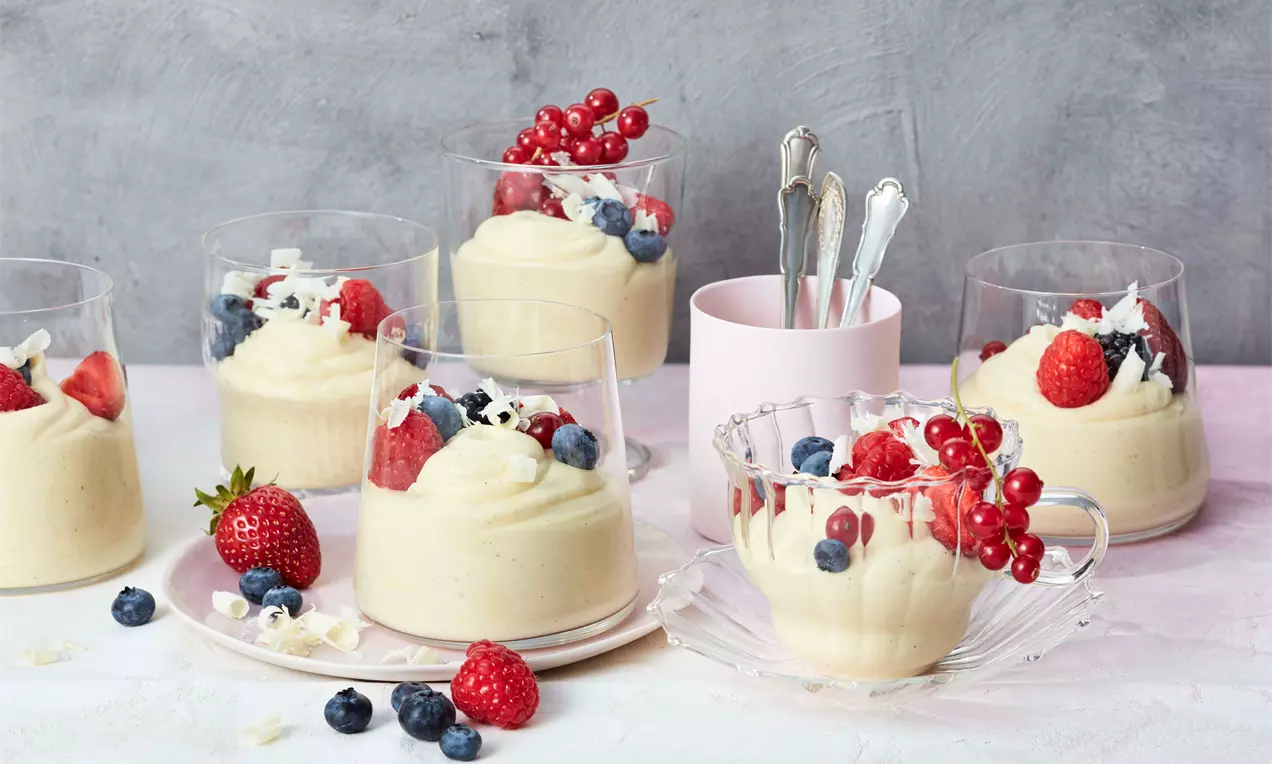 Weiße Mousse au Chocolat in transparenten Gläsern, cremig-fest mit Beeren, Schokospänen und frischen Früchten auf hellem Untergrund angerichtet