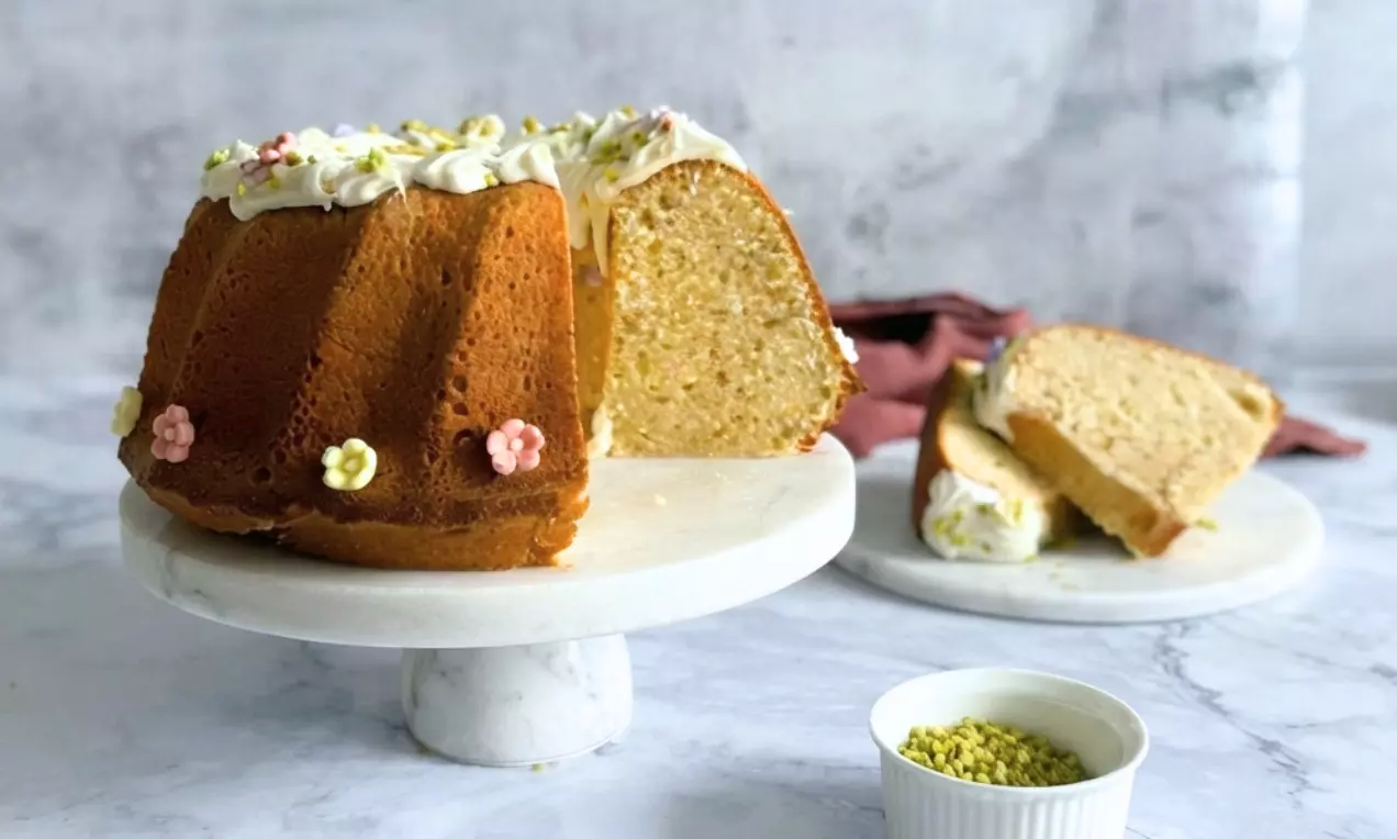 Goldbrauner Gugelhupf mit weißer Schokoglasur und Zuckerblümchen, teilweise angeschnitten auf einer Marmortortenplatte mit Pistazienschälchen