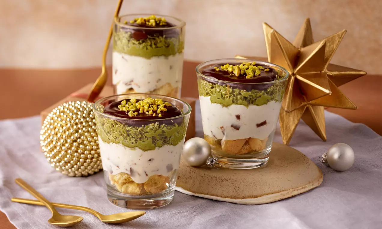 Schichtdessert in Gläsern mit grüner Pistazienschicht, weißer Creme, Schokoladensoße und dekorativem Topping auf hellem Untergrund drapiert