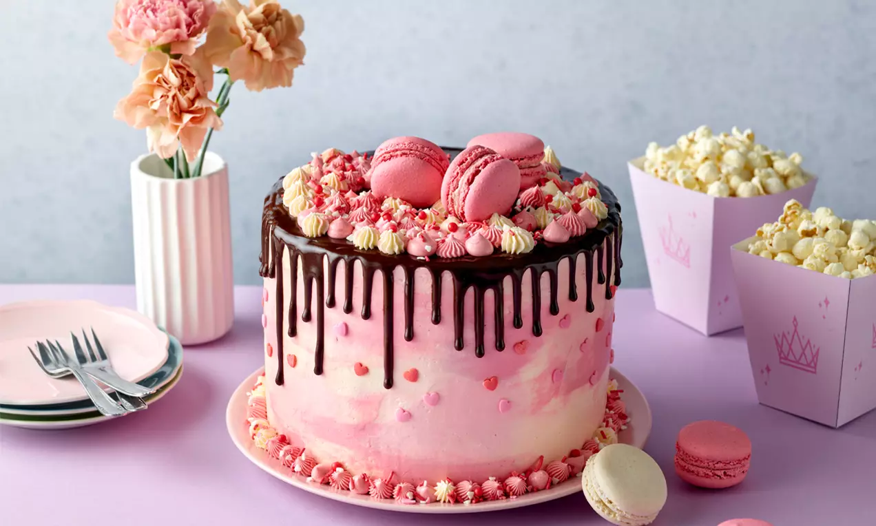 Rosa Geburtstagstorte mit Schokoglasur, cremigen Tupfen, Herzdekor, rosa Macarons und Popcorn in rosa Boxen auf lila Tischdecke