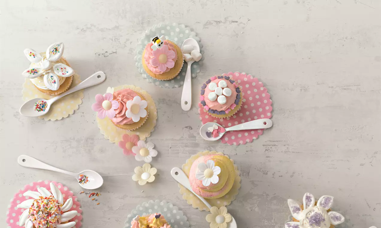 Bunte Cupcakes mit cremigen Blüten-Dekorationen auf pastellfarbenen Papieruntersetzern, flankiert von Löffeln mit Streuseln