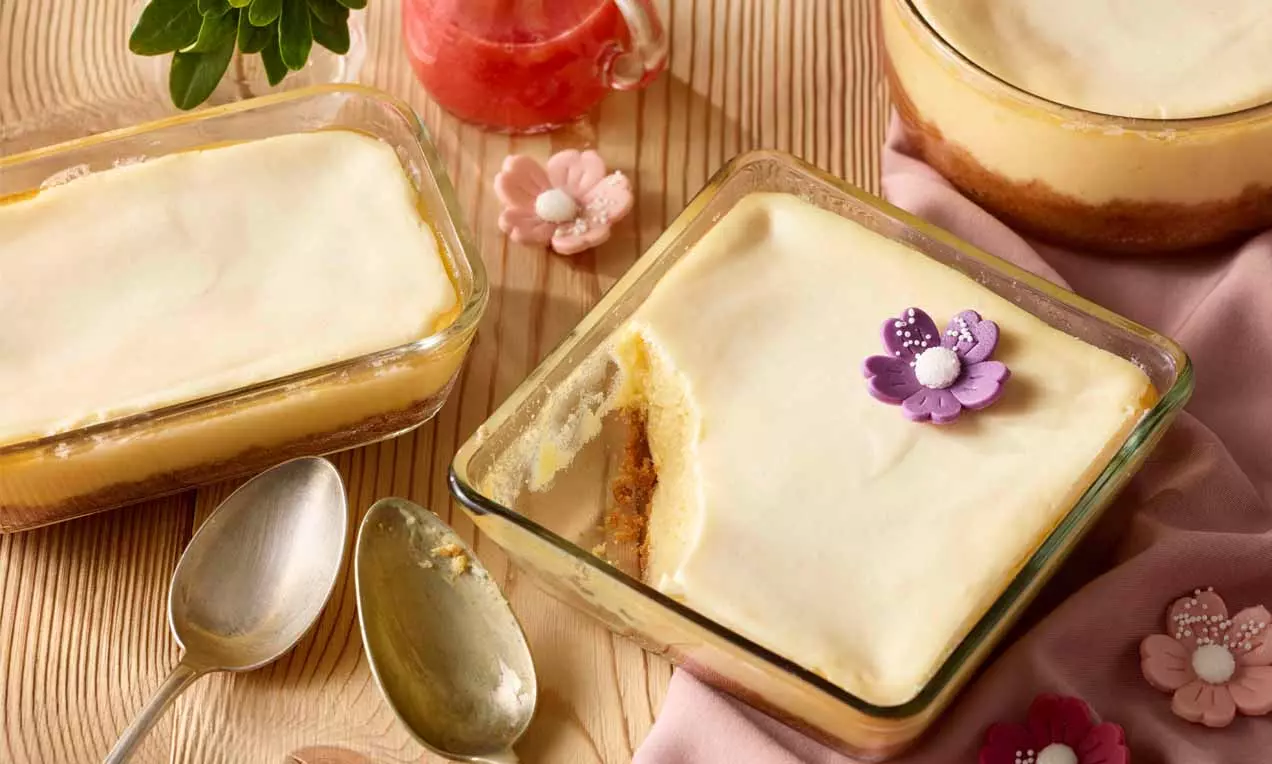 Kleine rechteckige Glasformen mit cremigem, hellgelbem New York Cheesecake, dekoriert mit bunten Blüten und umgeben von Löffeln, Stoff und Glas