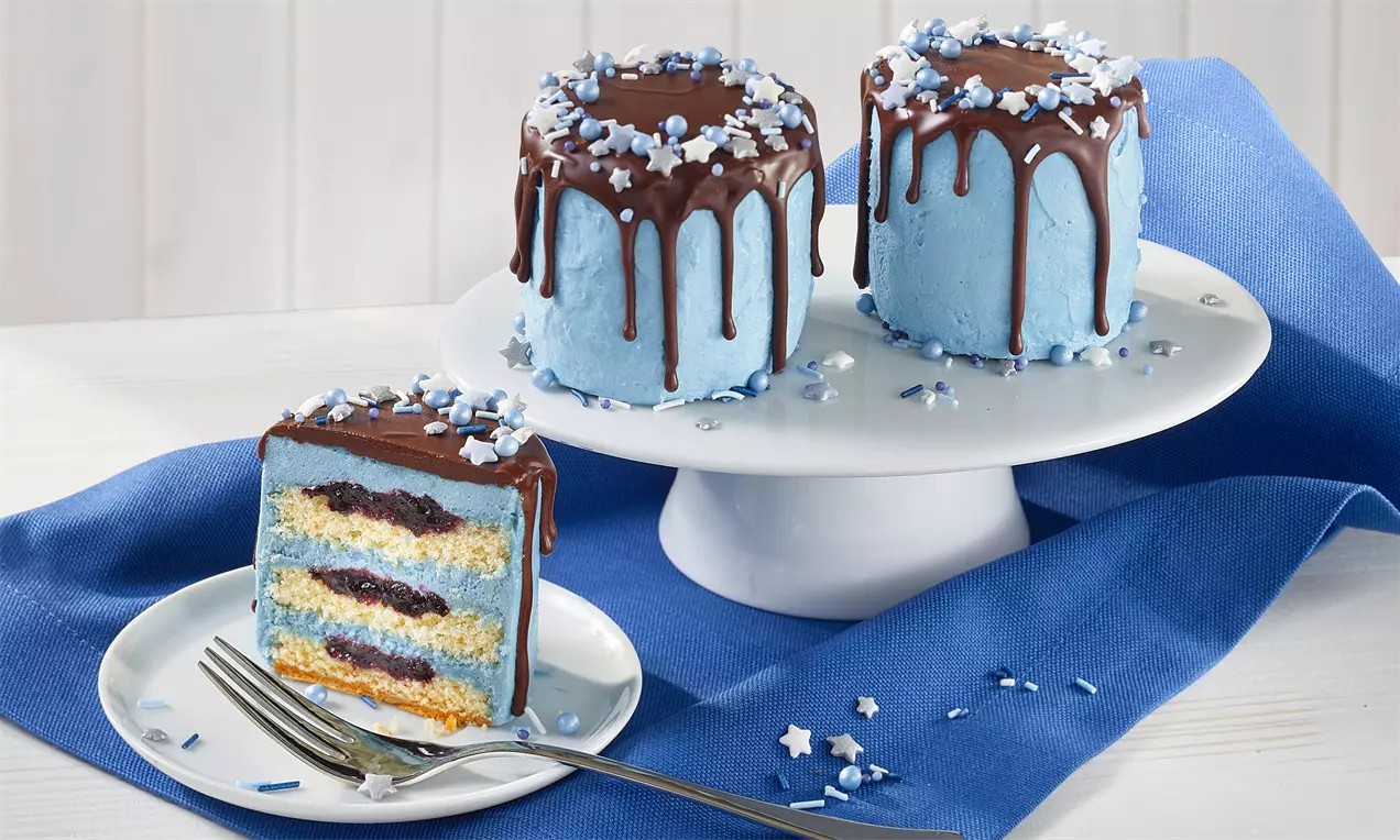 Kleine, himmelblaue Törtchen mit glänzendem Schokoladen-Drip und weißen Sternstreuseln auf einer weißen Tortenplatte mit blauem Stoff drapiert