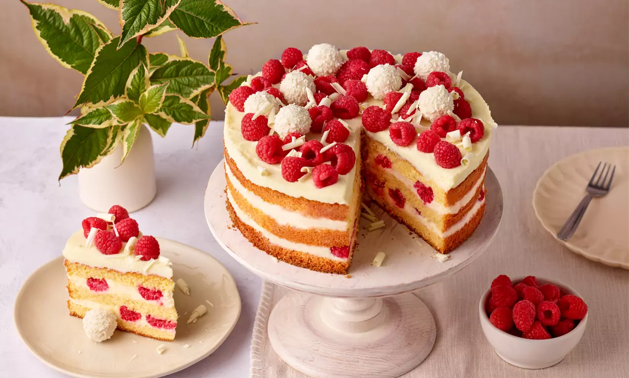 Weiße Torte mit Schichten aus heller Creme und Teig, dekoriert mit frischen Himbeeren, weißen Pralinen und Schokoraspeln auf einer Kuchenplatte