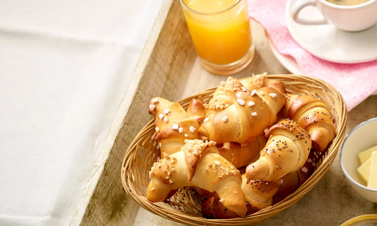 Kleine, goldbraune Hörnchen mit glänzender Oberfläche und feinen Körnchen, auf hellem Holztablett mit Orangensaft und Kaffee arrangiert