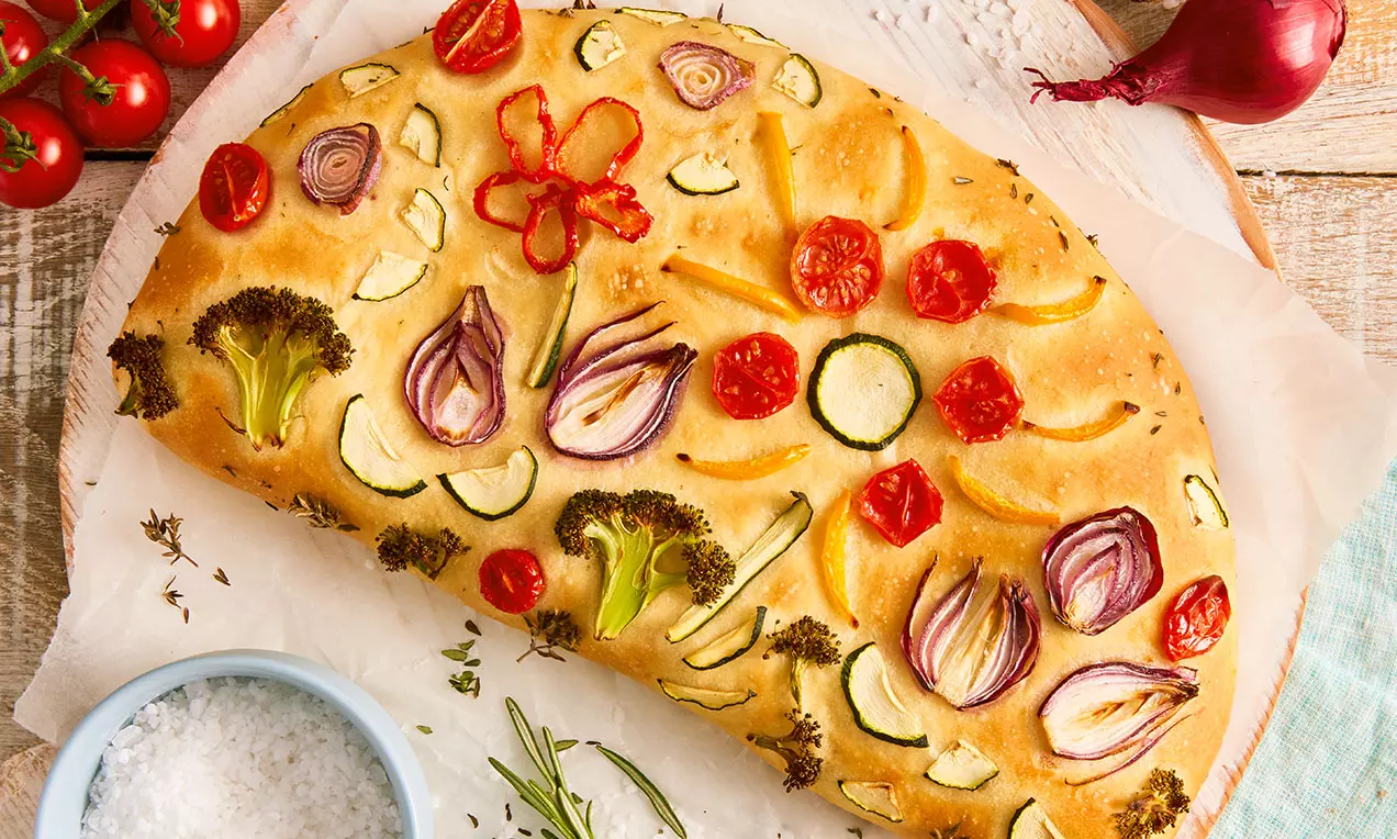 Goldbrauner Teig halbmondförmig mit buntem Gemüse-Muster aus Brokkoli, Zucchini, Tomaten und roten Zwiebeln auf Backpapier drapiert