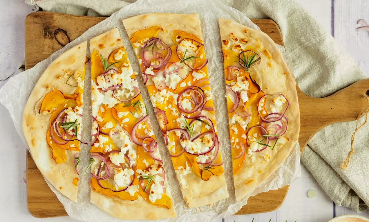 Flammkuchen mit dünnem, goldbraunem Teig, belegt mit orangefarbenem Kürbis, roten Zwiebelringen, Frischkäse und Rosmarin