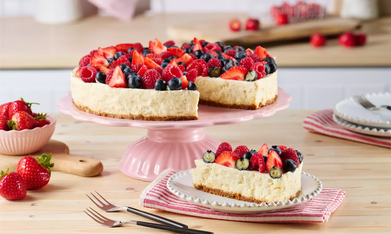 Eine runde Milchreistorte mit hellem Boden und cremiger Füllung, bedeckt mit roten Beeren und Blaubeeren auf einer rosa Kuchenplatte
