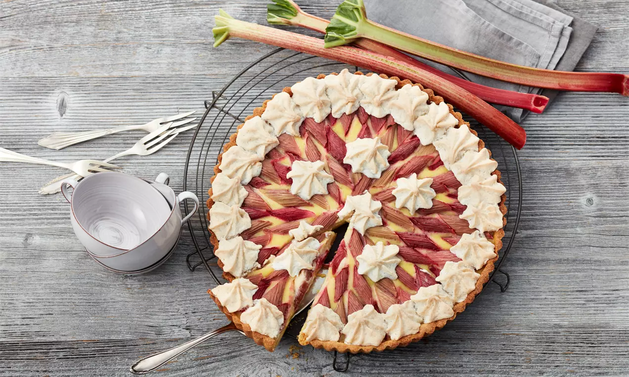 Rhabarber-Vanille-Tarte mit sternförmigen Baiserhauben auf einem Kuchengitter, daneben frische Rhabarberstangen und eine Tasse mit Untertasse