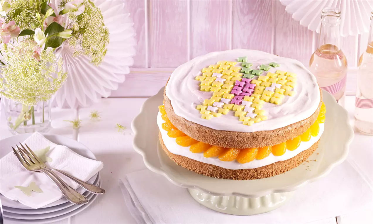 Runde Schichttorte auf pastellfarbener Tortenplatte mit heller Creme, Mandarinenstreifen und buntem Schmetterlingsmuster aus Zuckerdekor