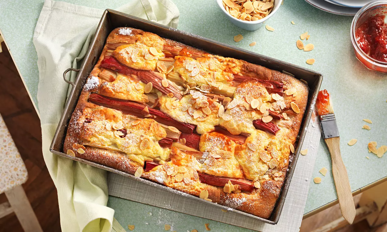Goldbrauner Rührteig mit roten Rhabarberstreifen, knusprigen Mandelblättchen und Puderzucker in einer eckigen Backform auf heller Oberfläche