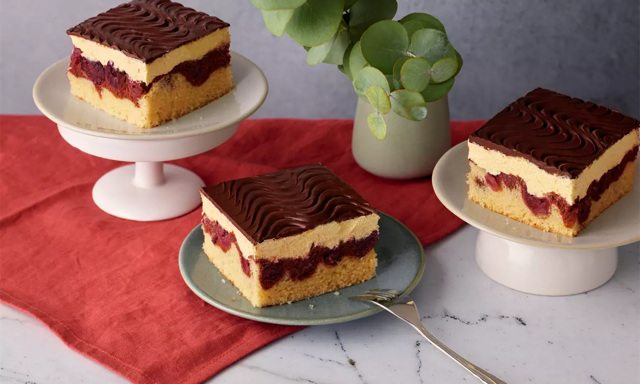 Veganer Donauwellenkuchen mit goldgelbem Boden, cremiger heller Füllung, roter Kirschschicht und wellenförmiger Schokoglasur auf Teller angerichtet
