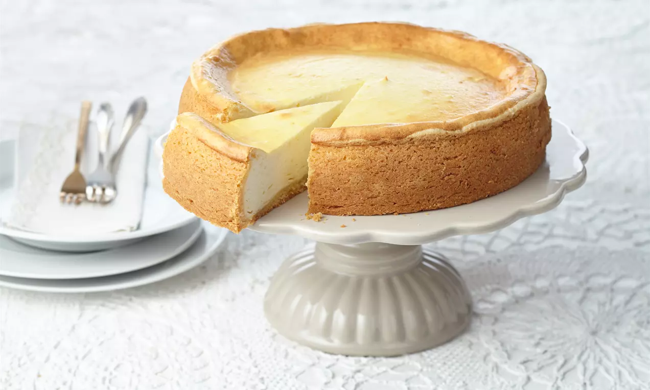 Goldbrauner Käsekuchen mit glatter, heller Oberfläche und einem Stück herausgeschnitten auf weißer Tortenplatte mit Teller und Besteck im Hintergrund