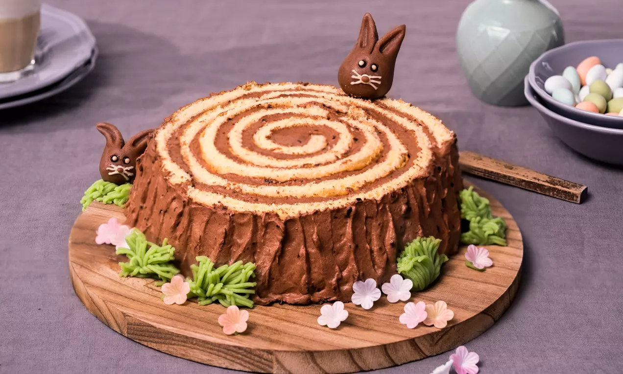 Schoko-Nougatstamm mit geschwungener Holzmaserung, verziert mit Marzipanhäschen, Blüten und grünem Gras auf einem Holzbrett