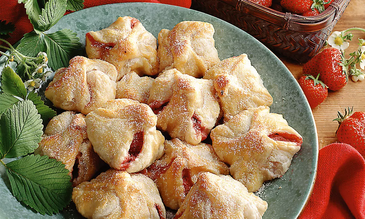 Goldbraune Blätterteigtaschen mit fruchtiger Erdbeerfüllung, bestäubt mit Zucker, auf einem grünen Teller mit Erdbeeren und Blättern angerichtet