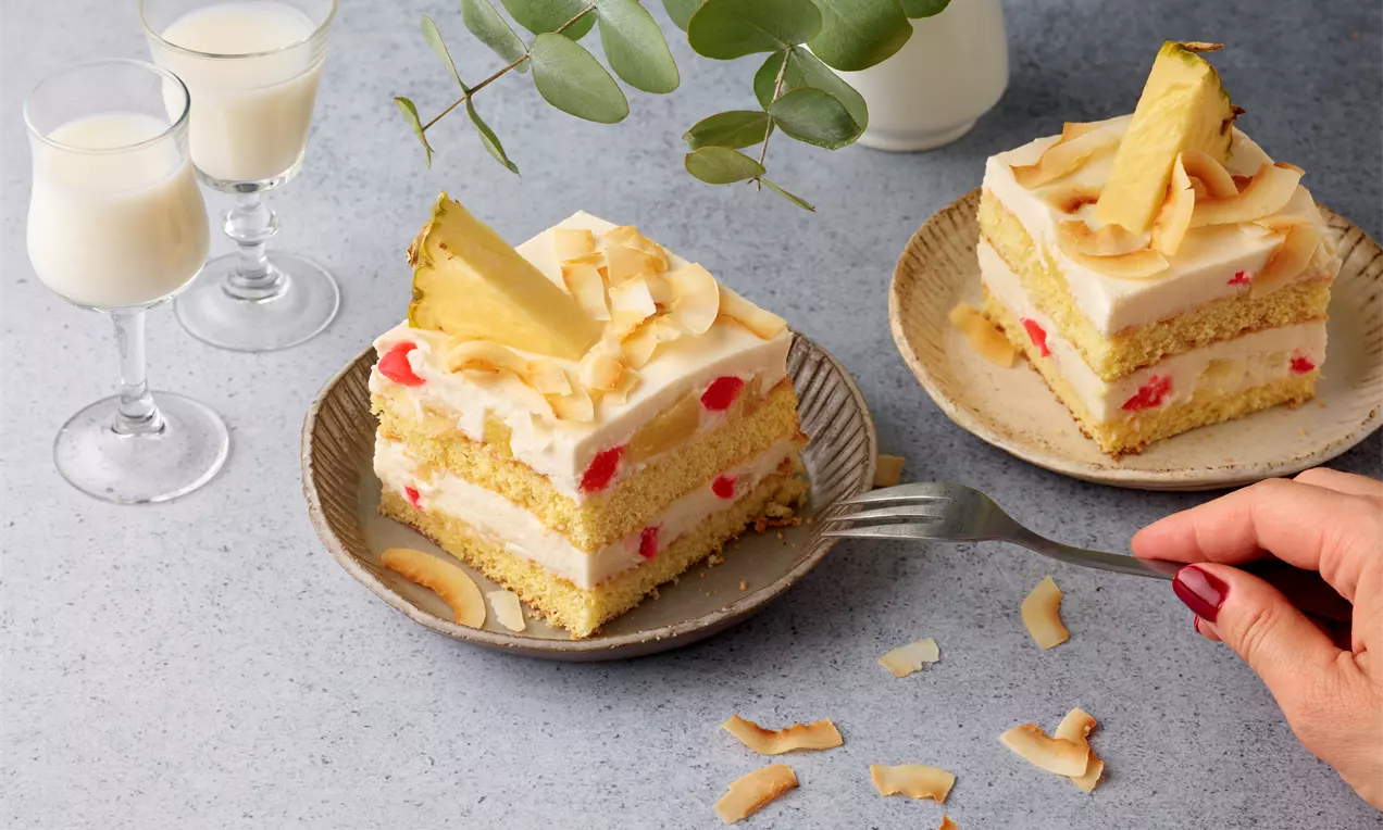 Pina-Colada-Schnitten mit gelber Creme, roten Fruchtstücken, gerösteten Kokoschips und Ananas auf kleinen Tellern, daneben Gläser mit Sahnelikör