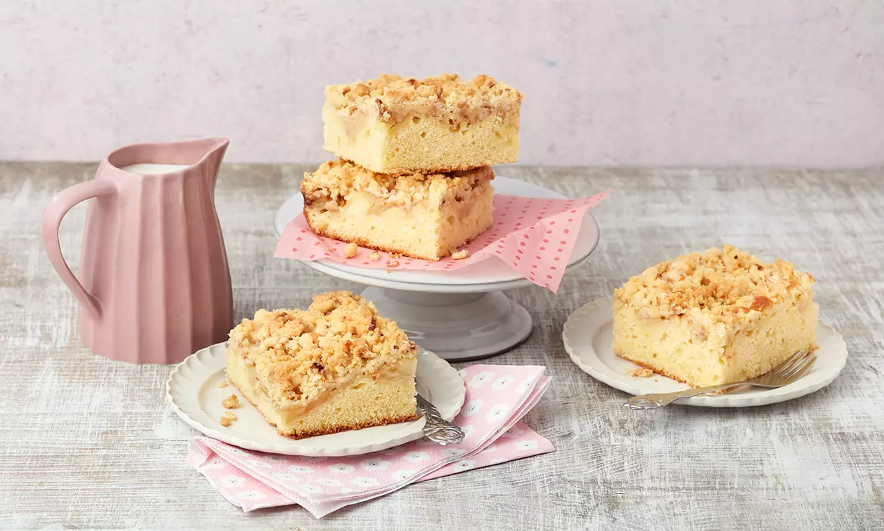 Zimtiger Apfel-Streuselkuchen mit goldbraunen Streuseln, hellgelbem Rührteig und rosa Servietten auf Tellern und einer weißen Tortenplatte