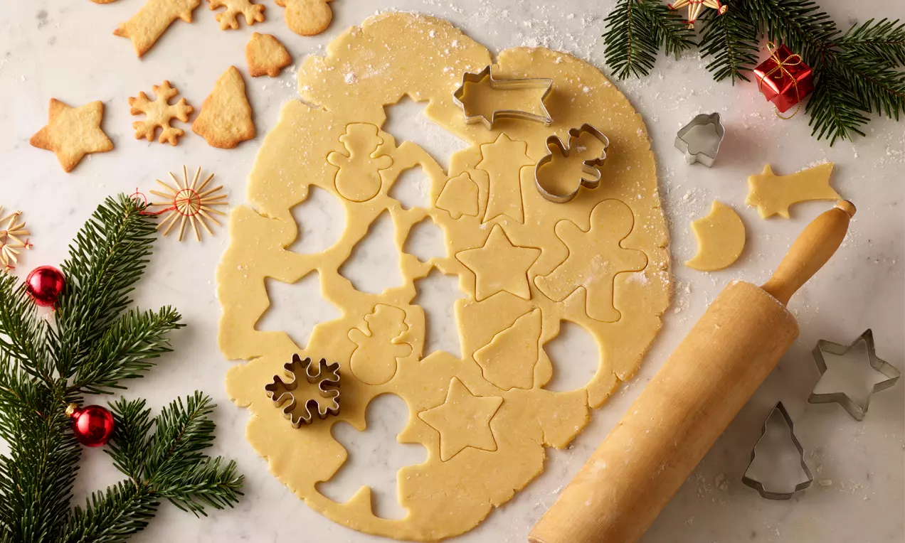 Ausgerollter, heller Mürbteig auf bemehlter Marmorplatte mit ausgestochenen Weihnachtsmotiven, Tannenzweigen und Nudelholz verziert
