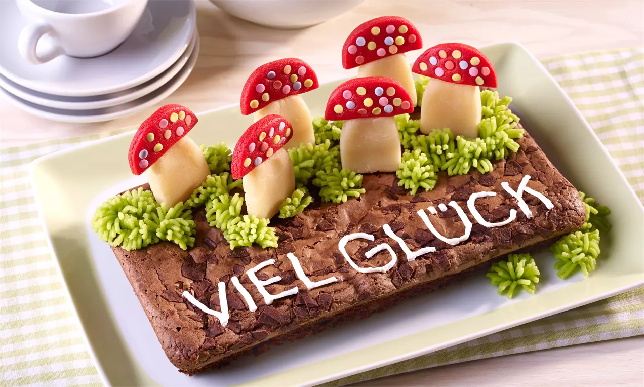Rechteckiger Schokoladenbrownie mit weißer Zuckerschrift, aus Marzipan geformte Fliegenpilze und grüne Cremetupfen auf einem hellen Teller