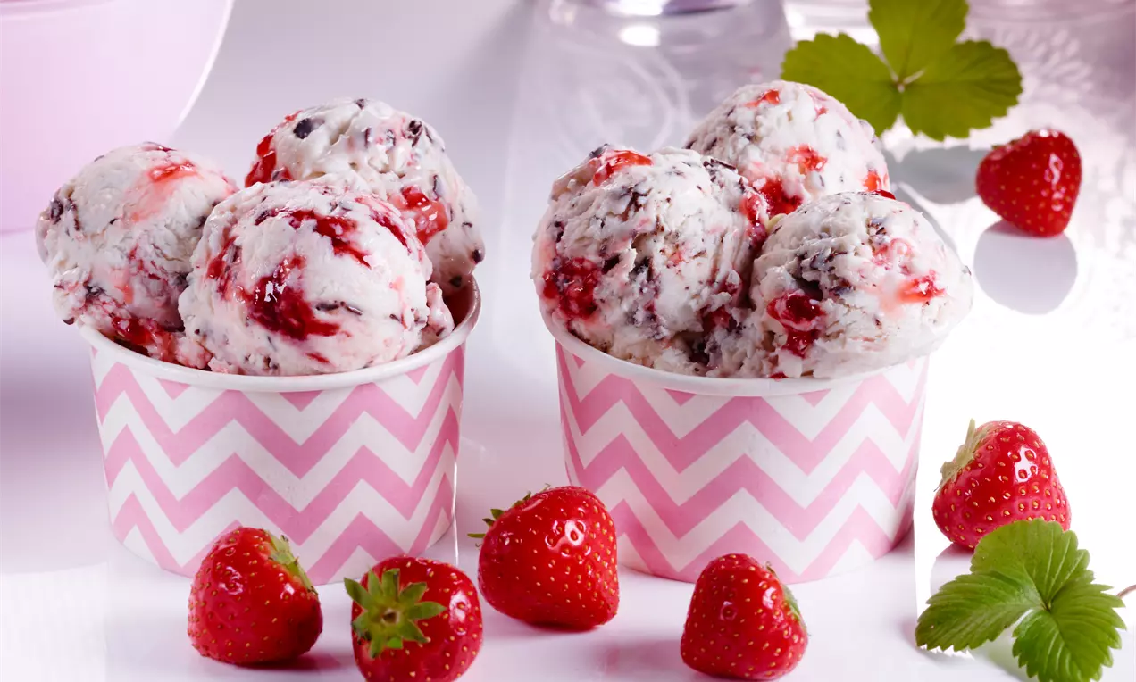 Stracciatella-Eiscreme mit roten Erdbeerstückchen in rosa-weißen Bechern mit Zickzackmuster, umgeben von frischen Erdbeeren und grünen Blättern