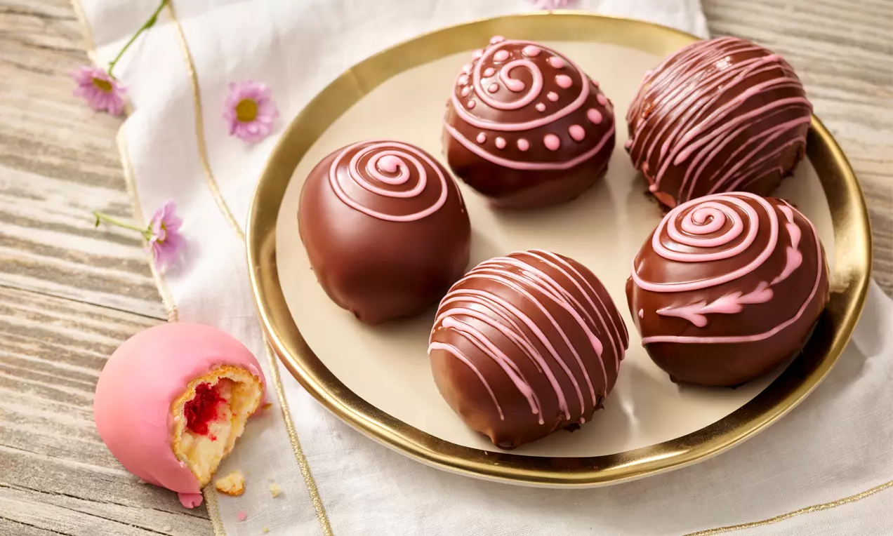 Runde Germteigbällchen mit glänzendem Schokoladenüberzug und rosafarbenen Zuckerverzierungen auf einem goldenen Teller mit weißen Blüten