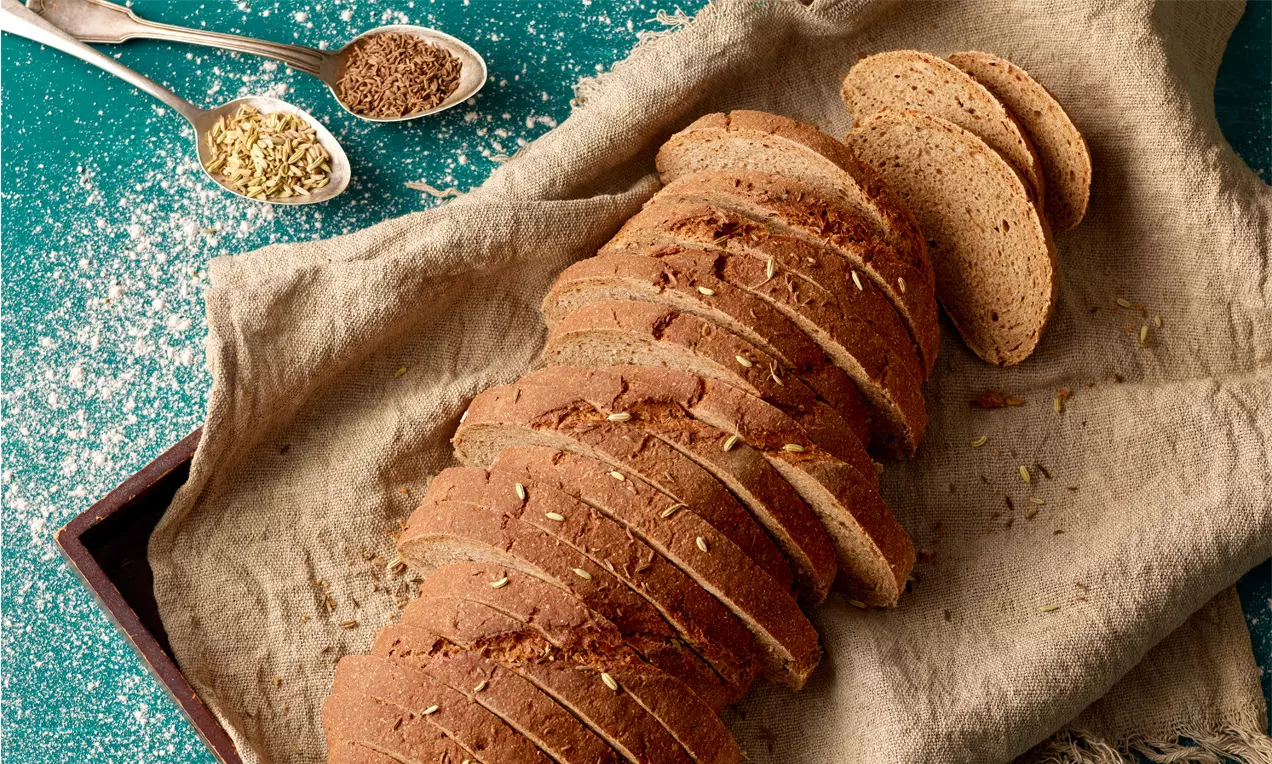 Dunkelbraunes, gleichmäßig aufgeschnittenes Bauernbrot mit goldener Kruste auf beigem Leinentuch, daneben Gewürze auf einem grünen Hintergrund