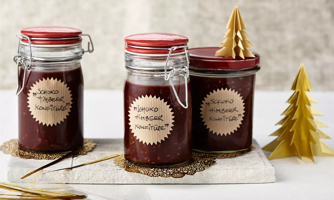 Drei Gläser mit dunkelroter Himbeerkonfitüre und roten Deckeln auf hellem Untergrund, dekoriert mit goldenen Papierbäumchen und Spitzendeckchen