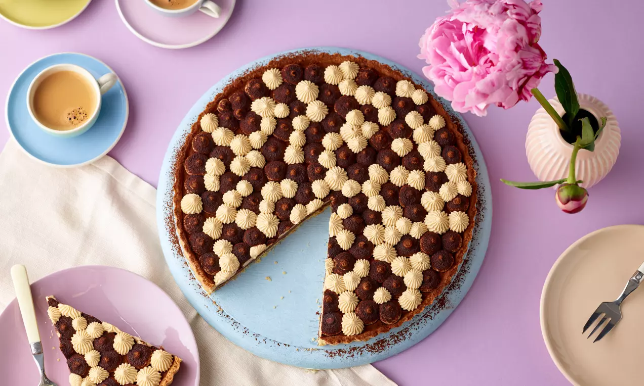 Tiramisu-Tarte mit dichtem Muster aus braunen Kakao- und beigen Creme-Tupfen auf hellem Teller, serviert mit einer angeschnittenen Portion