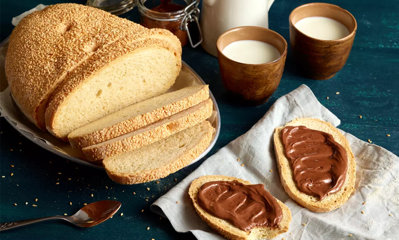 Weißbrot mit goldbrauner Kruste, in Scheiben geschnitten auf einem Teller mit zwei bestrichenen Brotscheiben und Milch in Holzbechern daneben