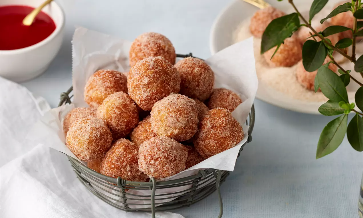 Rötlich-goldene, rundliche Topfenbällchen mit feiner Zuckerkruste in einem Drahtkorb mit weißem Papier auf einem hellen Tisch mit Deko
