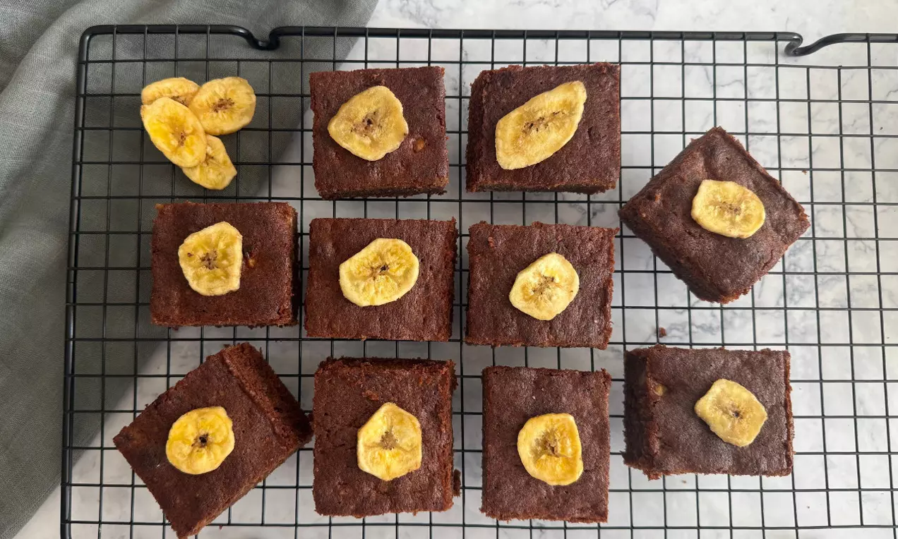 Saftige, quadratische Bananen-Brownies mit dunkelbrauner Oberfläche und goldgelben Bananenchips auf einem schwarzen Kuchengitter.