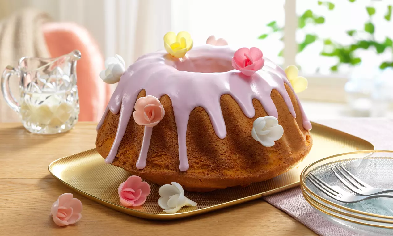 Goldbrauner Gugelhupf mit fliederfarbener Glasur, verziert mit bunten Zuckerblüten, auf einer goldenen Platte mit beigefarbenem Tuch drapiert