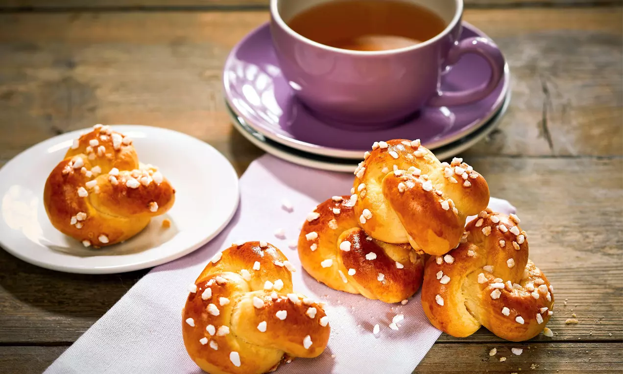 Goldbraune Germteig-Knöpflein mit grobem Hagelzucker bestreut auf weißem Tuch und Teller, daneben lilafarbene Teetasse auf Holzoptik-Tisch