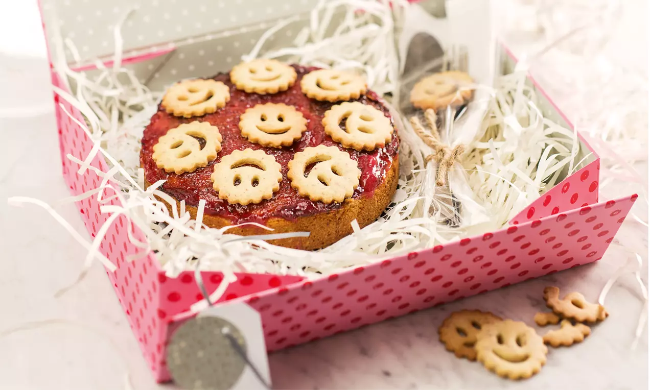 Weiße Mini-Linzer-Torte mit rotem Marmeladenbelag, goldbraunen Ausstechplätzchen und Dekopapier in einer gepunkteten Geschenkbox