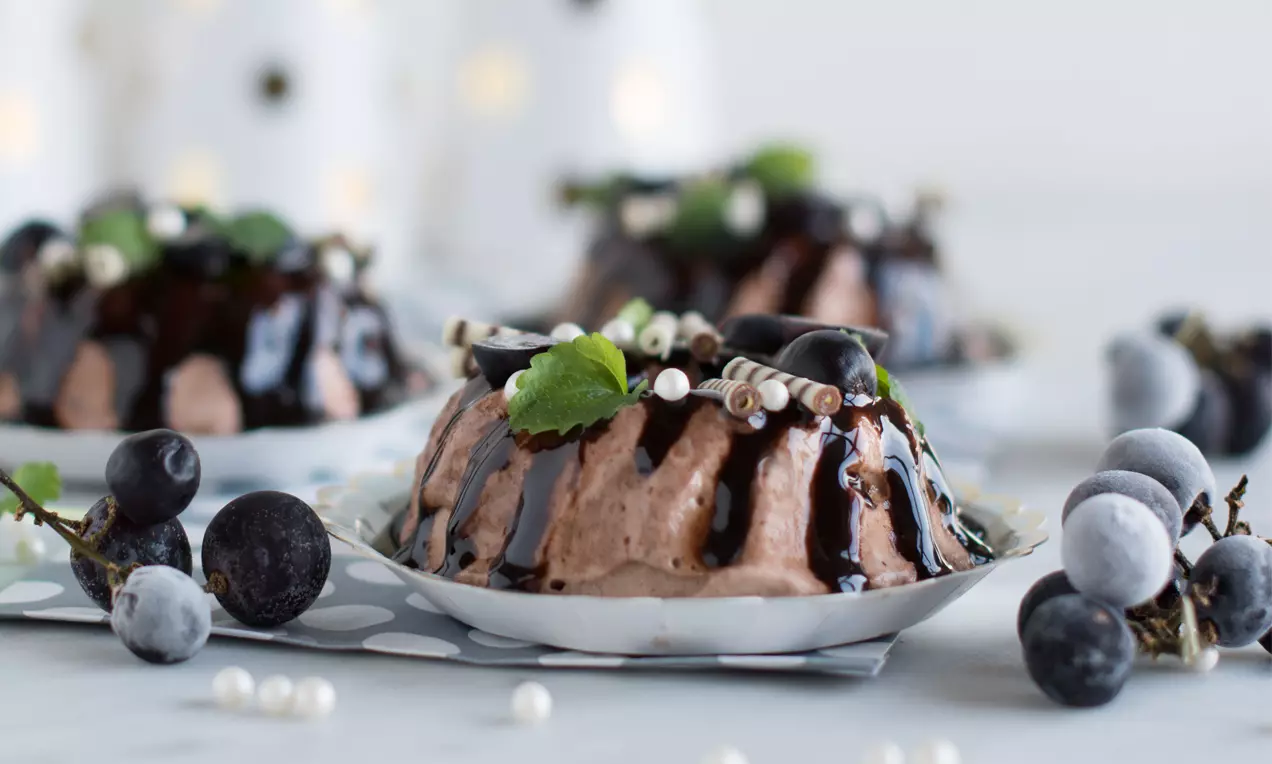 Schoko-Eis-Gugelhupf mit glänzender Schokosauce, Nüssen und Zimt dekoriert, auf weißem Teller mit Trauben und Minze angerichtet
