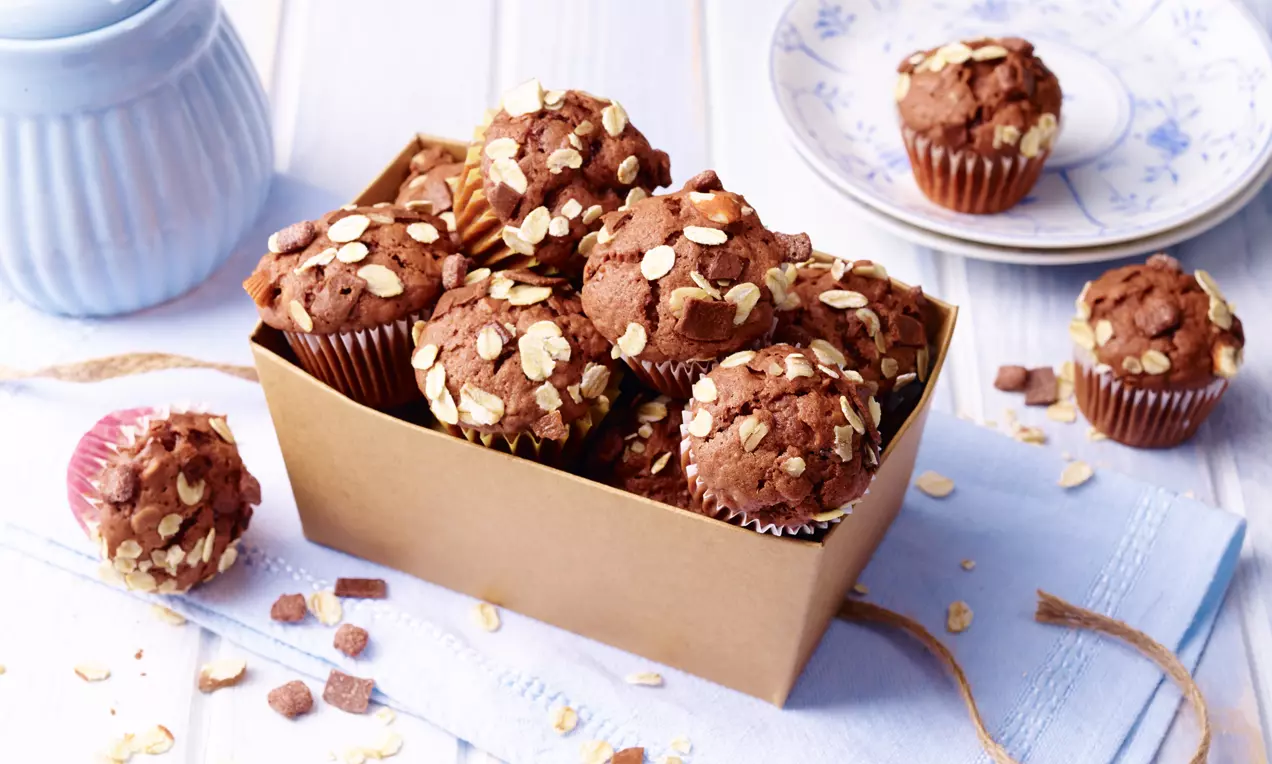 Schoko-Joghurt-Muffins mit brauner, lockerer Oberfläche, bestreut mit Haferflocken, in einer Pappschachtel auf hellblauem Stofftuch