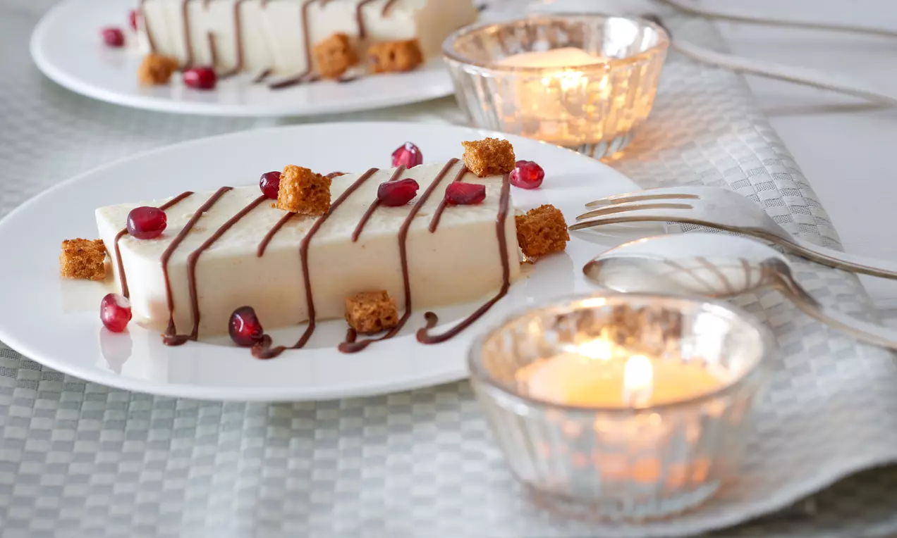 Ein rechteckiges Lebkuchen-Parfait mit heller, glatter Textur, dekoriert mit Schokoladensoße, Granatapfelkernen und Lebkuchenwürfeln auf weißem Teller