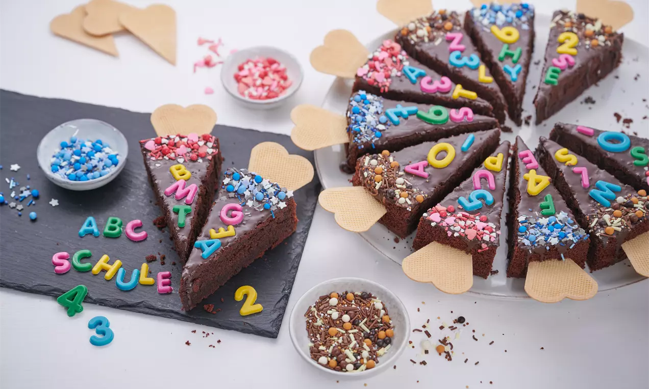 Dreieckige Kuchenstücke aus dunklem Teig mit bunten Zuckerperlen und Buchstaben dekoriert, an den Seiten mit Waffelherzen verziert