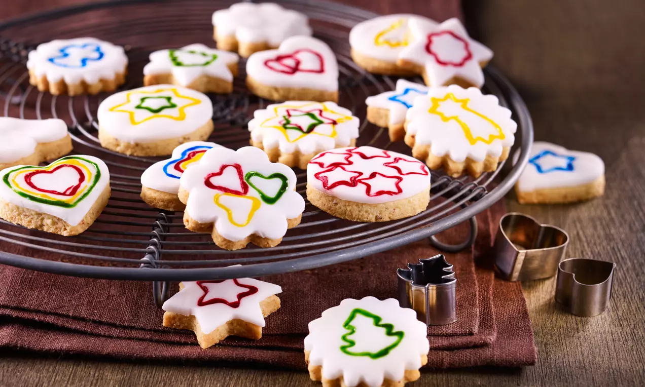 Runde und blumenförmige Plätzchen mit weißem Fondant und bunten, gestempelten Motiven liegen auf einem runden Kuchengitter
