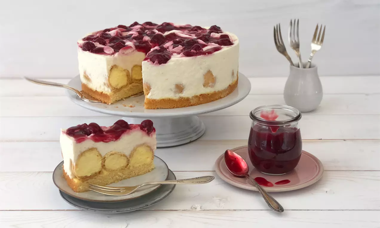 Windbeuteltorte mit luftiger Sahnecreme, eingebetteten Windbeuteln und glänzender Kirsch-Grütze auf weißer Tortenplatte mit Gabeln und Kirschglas