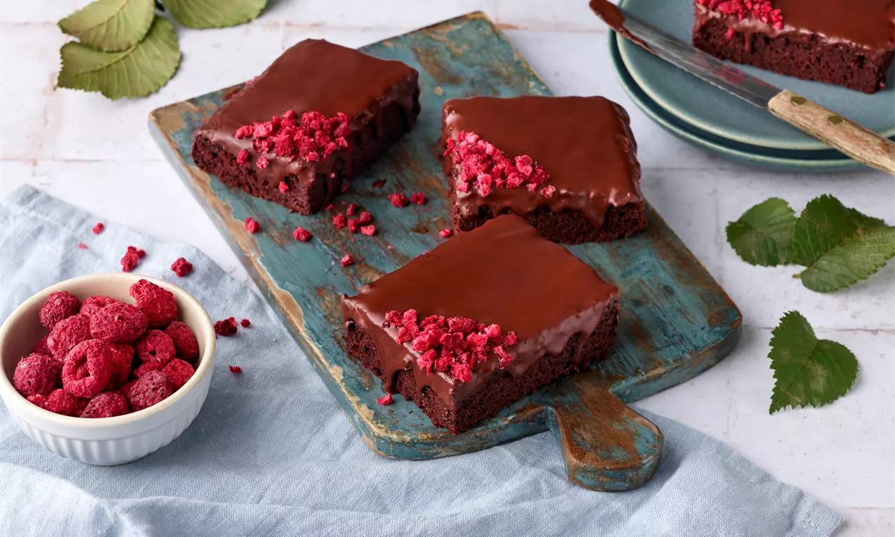Schokoladige Red-Velvet-Kuchenstücke mit glänzender Glasur und roten Himbeerkrümeln auf rustikalem Holzbrett mit frischen Himbeeren